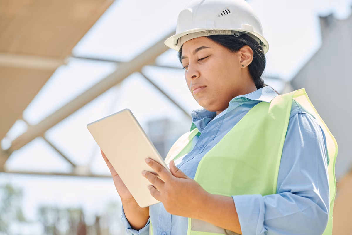 A construction worker in a white hard hat and high-visibility vest uses a tablet computer outdoors beneath a steel-framed structure.